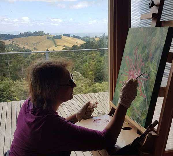 Katherine painting a red grevillea at her easel, looking out over green hills from her studio